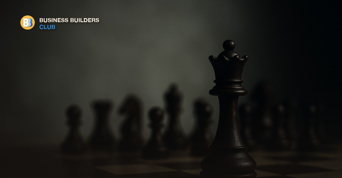 A chess queen piece standing alone on a clean white surface with blurred pieces in the background